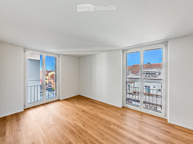 Empty room with light wooden flooring and white walls, featuring large sliding glass doors that open to a balcony with a view of rooftops.