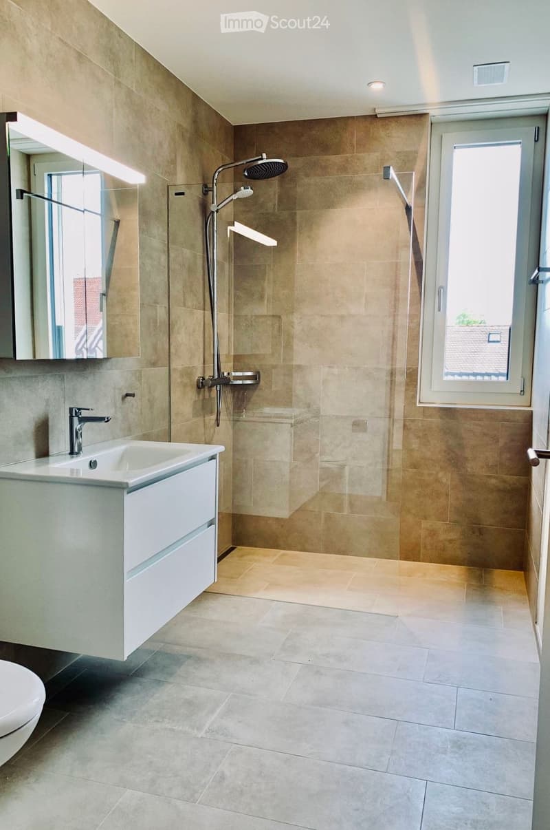 Modern bathroom with beige tiles, a white sink with a chrome faucet, a large mirror, a toilet, and a walk-in shower with a glass enclosure and multiple showerheads.