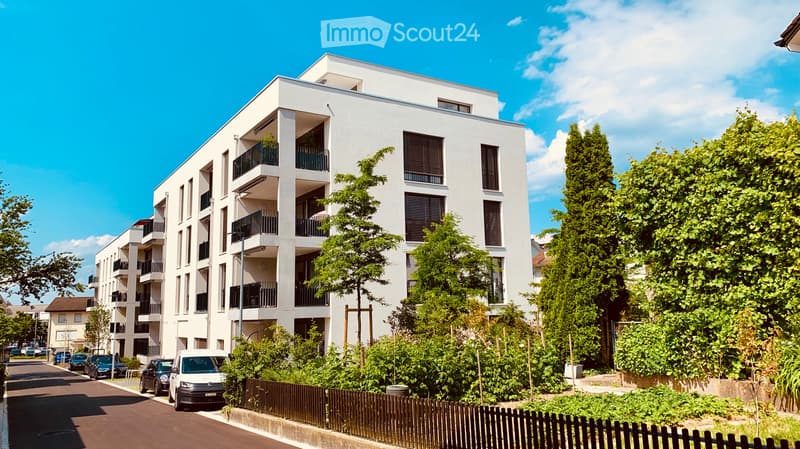 Modern multi-story residential building with white facade, balconies, and surrounding greenery.