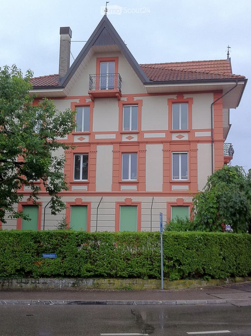 Two-story house with red and white exterior, multiple windows, green garage doors, and a small balcony on the upper floor.