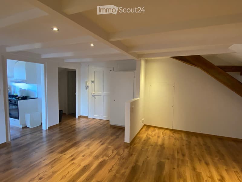 Empty room with wooden flooring, white walls, and recessed lighting. Features a staircase leading upwards and an open doorway to a kitchen area.