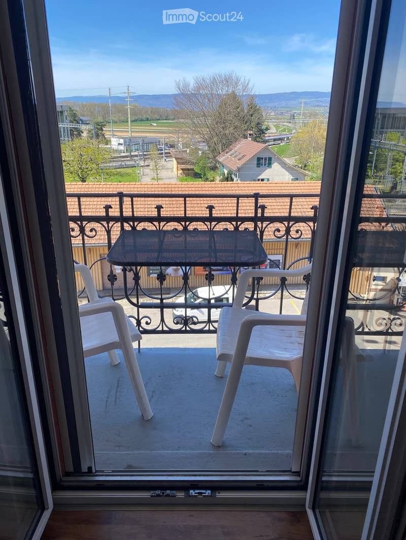 Balcony with metal railing, table, and two chairs, overlooking a scenic view of a town and countryside.