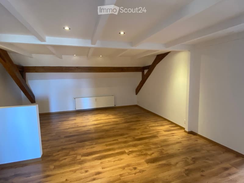 Empty room with wooden flooring, white walls, exposed wooden beams, and recessed lighting. A radiator is visible on one wall.