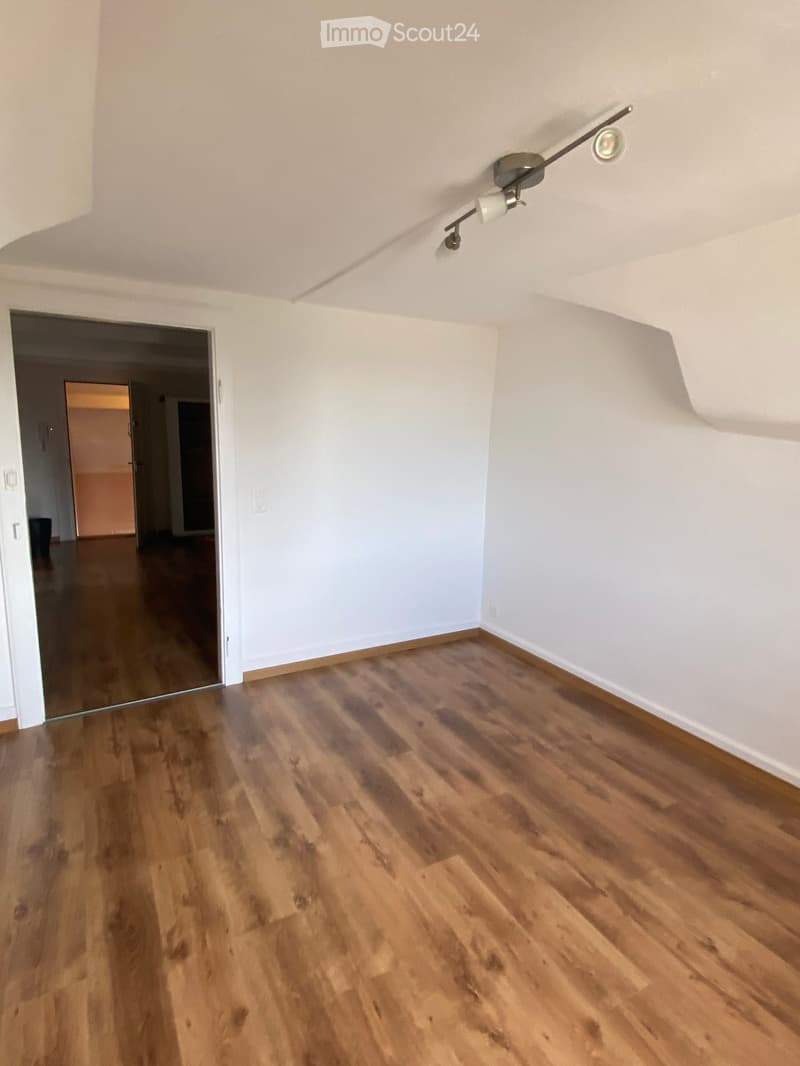 Empty room with wooden flooring, white walls, and a ceiling light fixture. The room has an open doorway leading to another area.