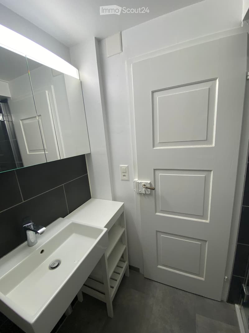 Modern bathroom with a white sink, chrome faucet, large mirror, and white cabinet. The walls are white with black tiles below the mirror.