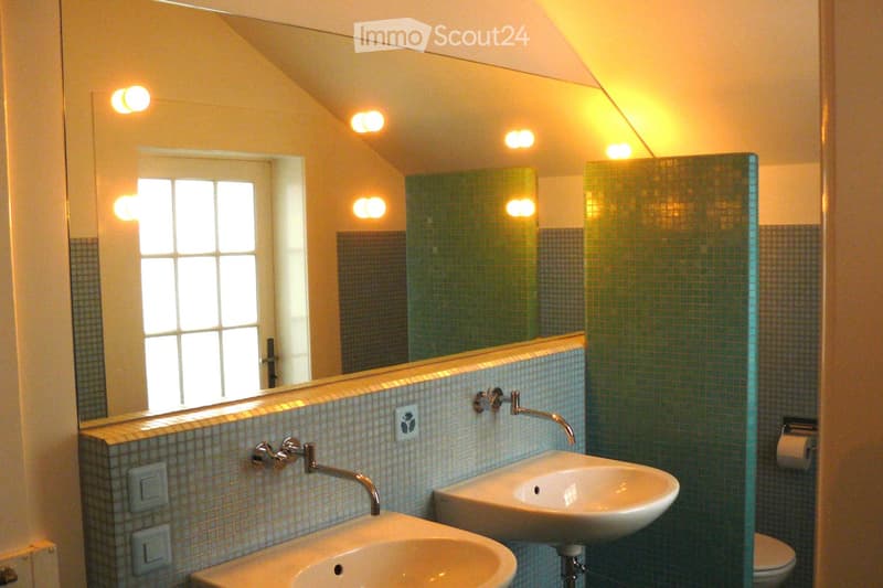 Modern bathroom with a large mirror, two sinks, a toilet, and a glass shower enclosure. The walls are tiled with small, light-colored tiles, and the ceiling has recessed lighting.
