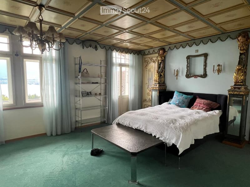 Elegant bedroom with a large bed, bench, ornate mirror, shelf, chandelier, and a view through large windows.