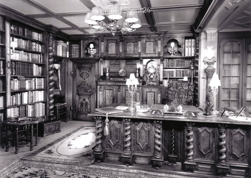 Ornate wooden study with large bookshelves, a large wooden desk, a round table, and a patterned rug.