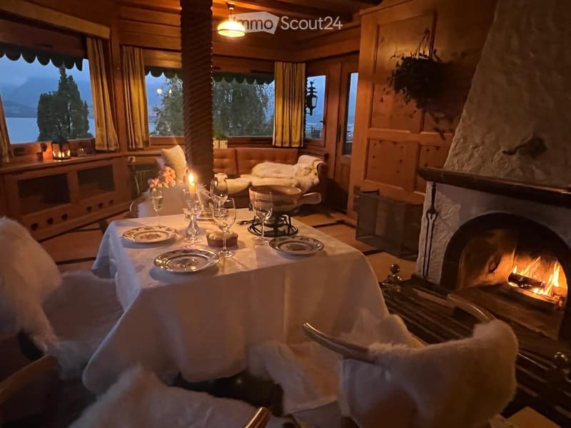 Cozy dining area with a round table set for dinner, surrounded by plush armchairs and a stone fireplace with a burning fire. Large windows offer a scenic view of a lake and mountains.