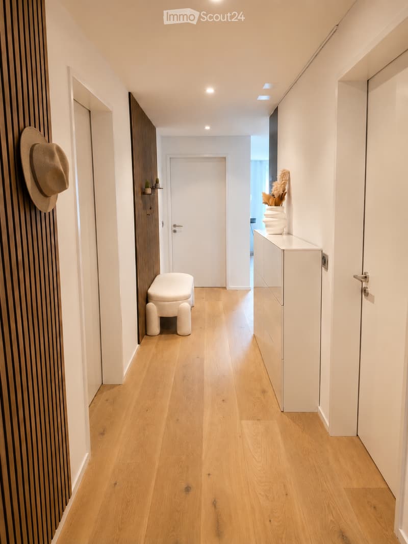 Modern hallway with light wooden flooring, white walls, and recessed lighting. Features a small bench, a hat on a wall-mounted hook, and a small cabinet with decorative items on top.