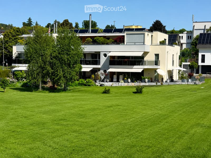 Modern two-story house with large windows, balconies, and solar panels on the roof, surrounded by a well-maintained lawn and trees.