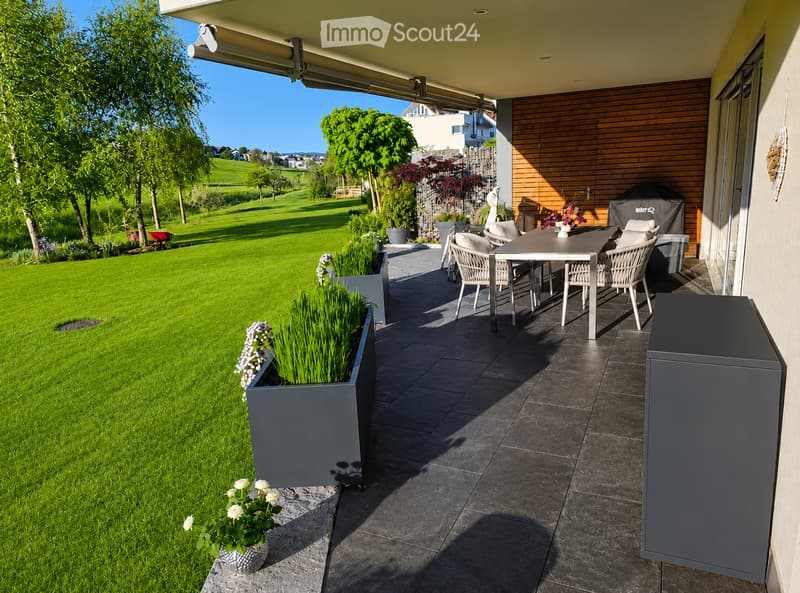 Outdoor patio with a dining table and chairs, surrounded by a well-maintained lawn and garden, with a view of a green field and trees.