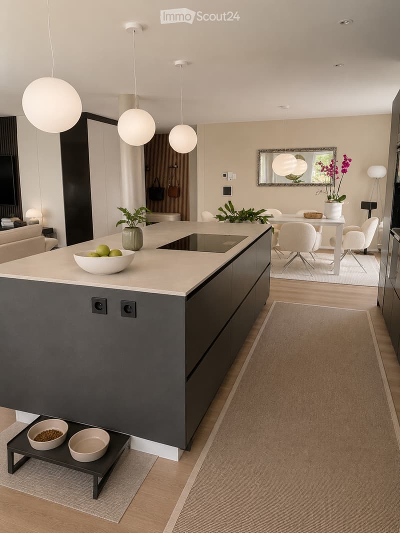 Modern kitchen with a central island, pendant lights, and a dining area with chairs and a table. There are decorative bowls, a vase with plants, a mirror, and a small sofa in the background.