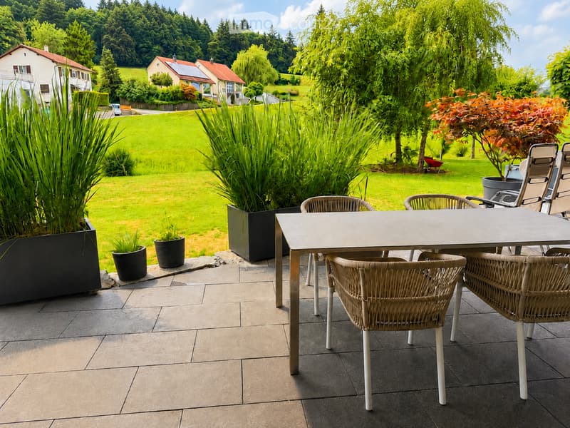 A well-maintained garden with a patio area featuring a table and chairs, surrounded by greenery and potted plants, with a scenic view of a countryside landscape.