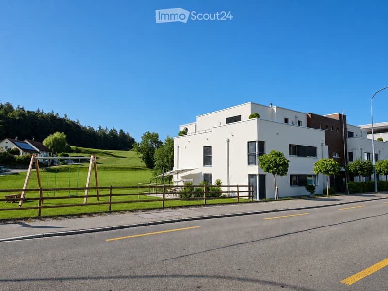 Modern white building with multiple windows and balconies, surrounded by greenery and a playground area.