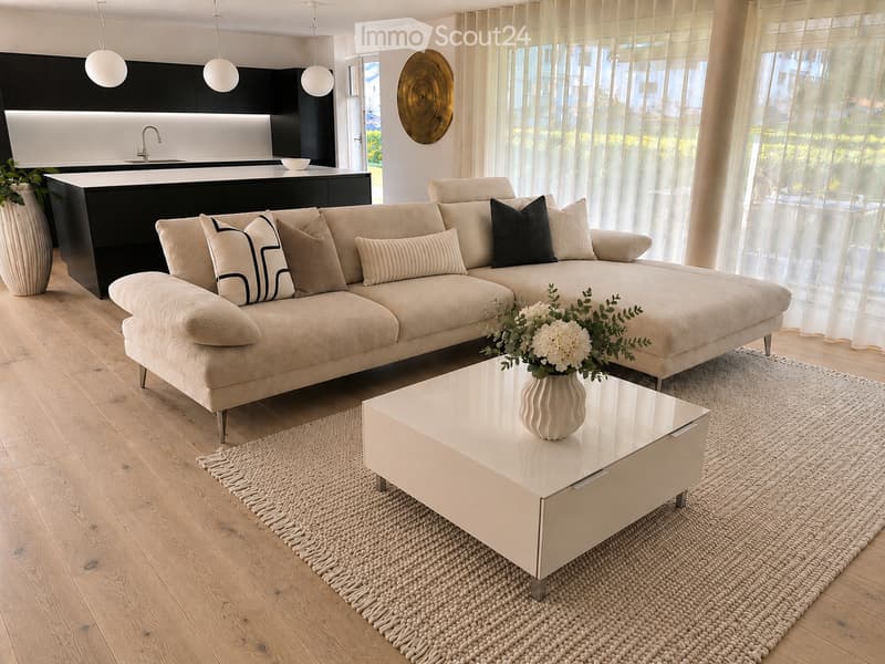 Modern living room with a beige sectional sofa, white coffee table, and a vase with white flowers. The room features a kitchen area with a black island, a sink, and a bowl on the counter. The space is illuminated by pendant lights and has large windows wi