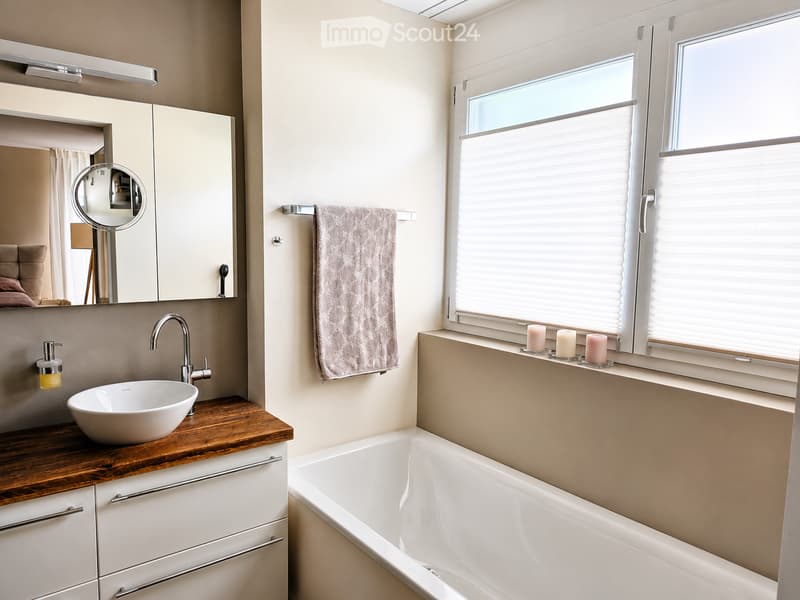 Modern bathroom with a white sink, wooden countertop, bathtub, large mirror, and a towel hanging on a rack. There are candles on the windowsill and a window with blinds.