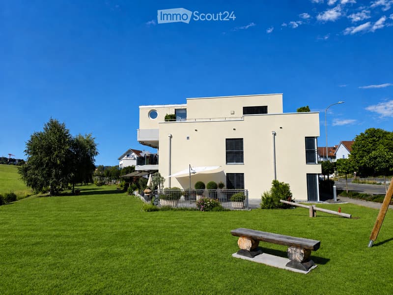 Modern two-story house with light beige facade, large windows, and a balcony. The front yard features a wooden bench, a small patio with outdoor furniture, and a well-maintained lawn.