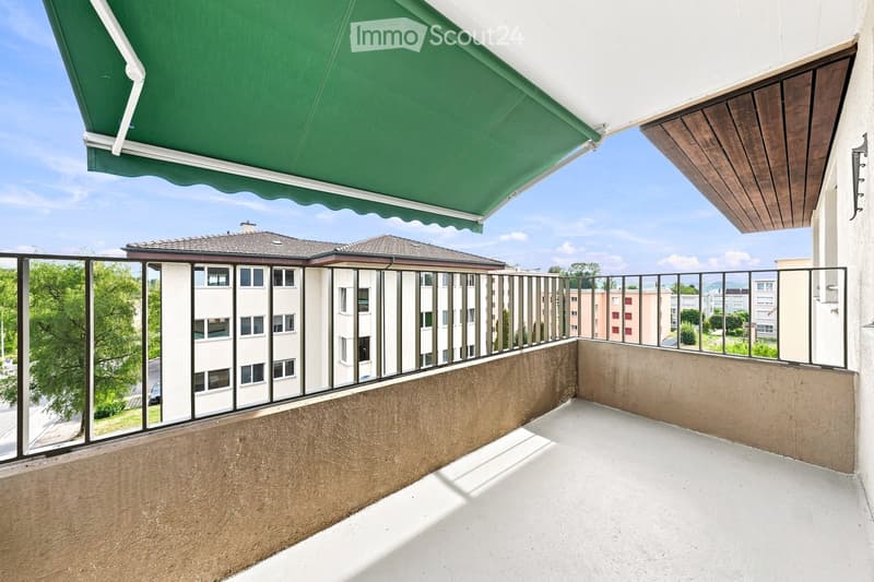Balcony with a green awning, metal railing, and a view of a residential area with buildings and trees.