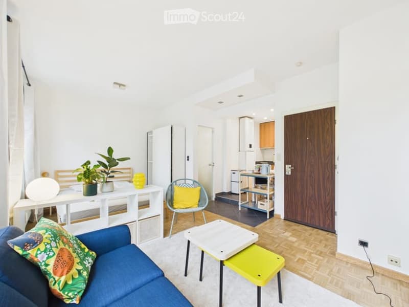 Bright, modern living space with a blue sofa, white coffee table, yellow accent chair, small kitchen area with refrigerator and microwave, and decorative plants.