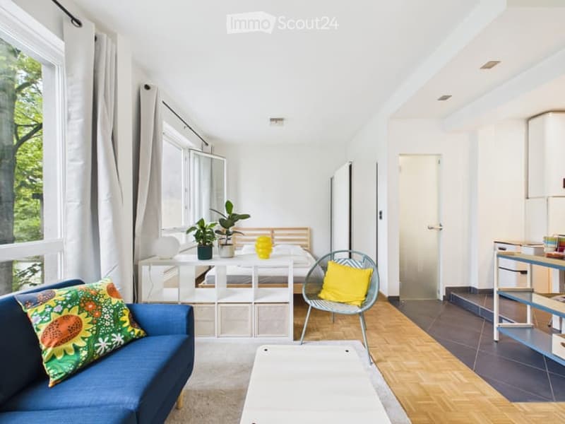 Bright, airy room with large windows offering a view of greenery. Features a blue sofa, a white table, a bed, a chair with a yellow pillow, and several potted plants.