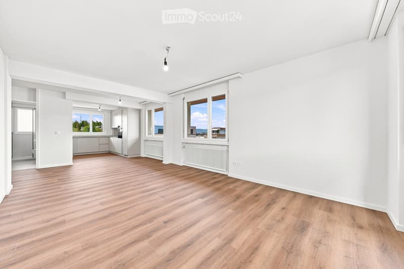 Bright, unfurnished room with wooden flooring, white walls, large windows, and a view of the outdoors. The room is connected to a small kitchen area with white cabinets and a countertop.