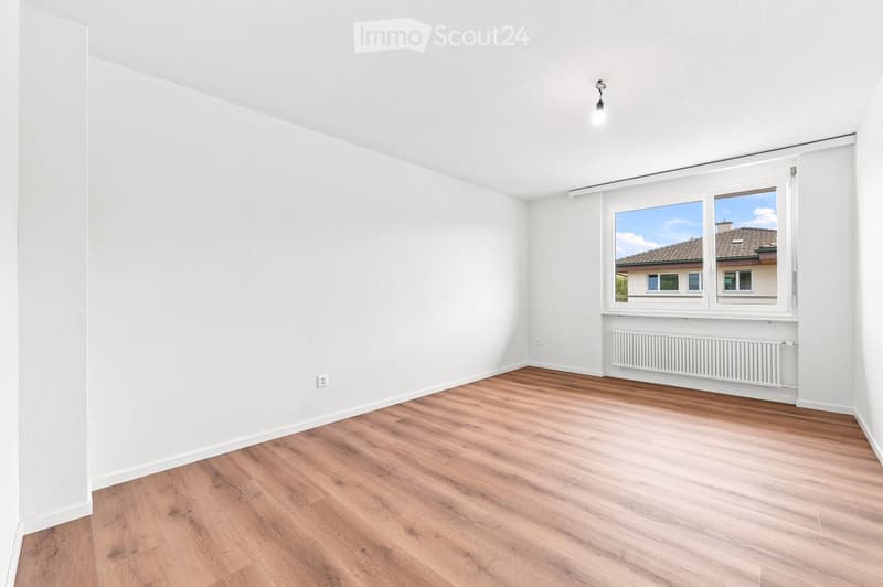 Empty room with white walls, wooden flooring, and a large window with a view of neighboring buildings.