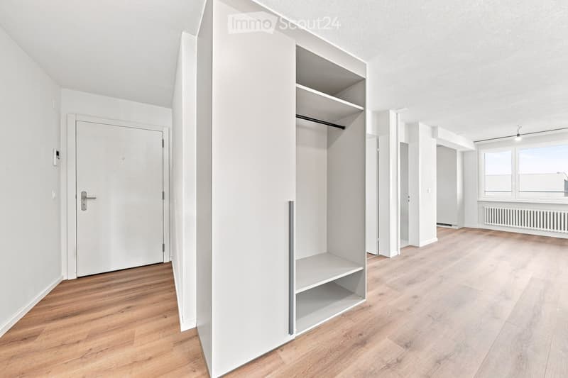 Empty room with light wooden flooring, white walls, built-in shelving unit, and large windows allowing natural light.