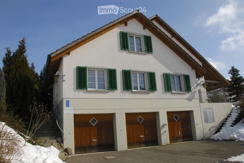 Two-story house with white walls, green shutters, wooden garage doors, and a snow-covered driveway.