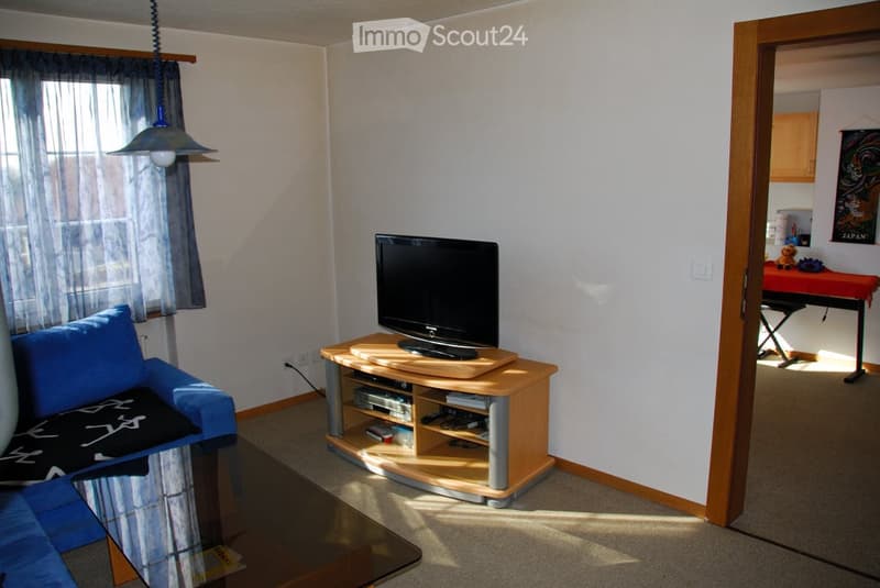 Living room with a blue sofa, a TV on a wooden stand, a glass coffee table, and a small table with a red cloth in the adjacent room.