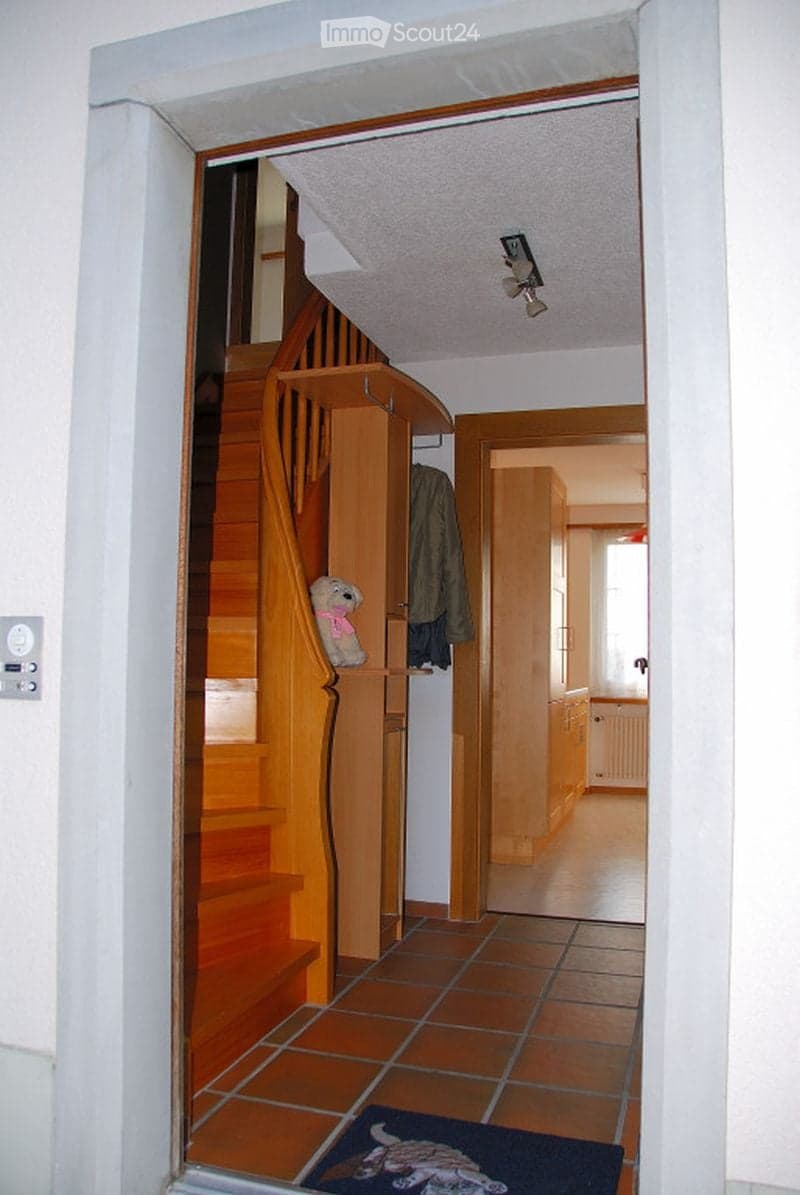 A hallway with a wooden staircase, tiled floor, and a small rug near the entrance. A coat is hanging on the wall, and a stuffed animal is placed on the staircase railing.