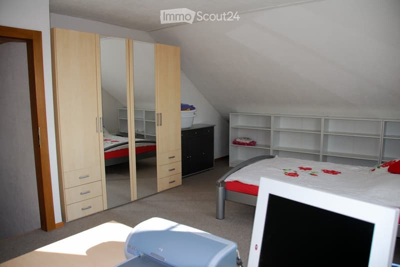 Bright bedroom with slanted ceilings, featuring a large mirrored wardrobe, a double bed with red and white bedding, shelves, and a laptop on a desk.