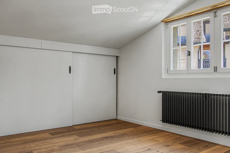 Empty attic room with wooden flooring, white walls, large windows, and a radiator. No furniture or personal items present.