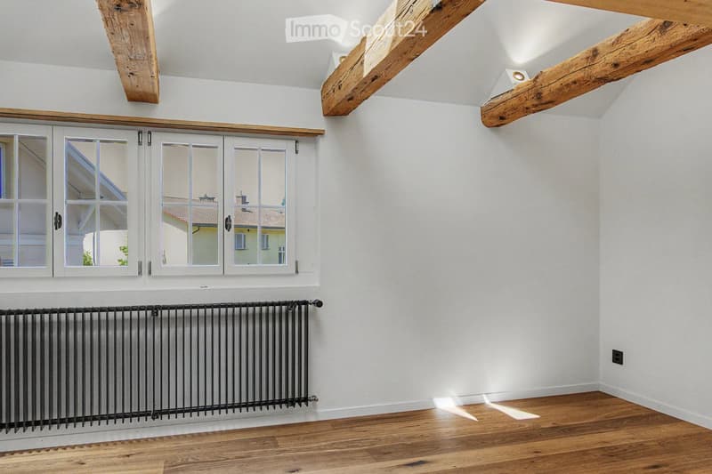 Empty room with wooden flooring, white walls, exposed wooden beams on the ceiling, and a large window with white frames. A black radiator is mounted on the wall below the window.