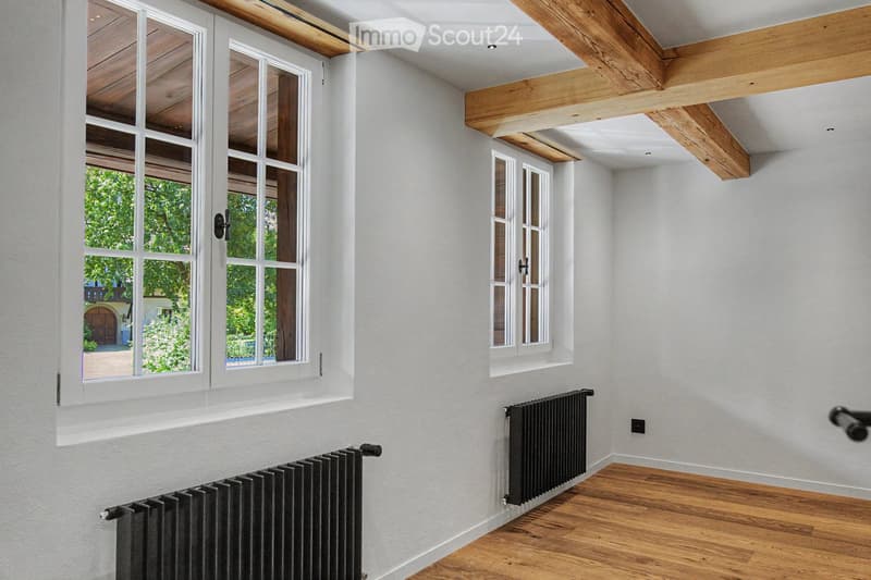 Empty room with light grey walls, wooden floor, and exposed wooden beams on the ceiling. Two windows with white frames and black radiators are visible.