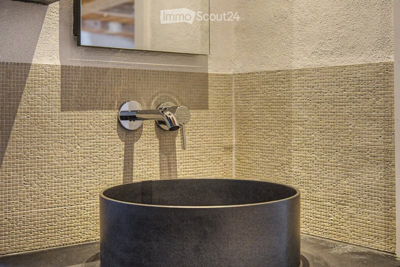 Modern bathroom with a sleek, dark sink and a stylish chrome faucet mounted on a textured beige tiled wall. A mirror is mounted above the sink.