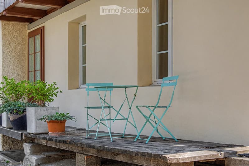 A wooden balcony with a light blue metal table and chair set, surrounded by potted plants and a view of the house exterior.