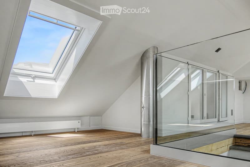 Bright, empty attic room with wooden flooring, white walls, skylight windows, and a glass staircase leading to another level.