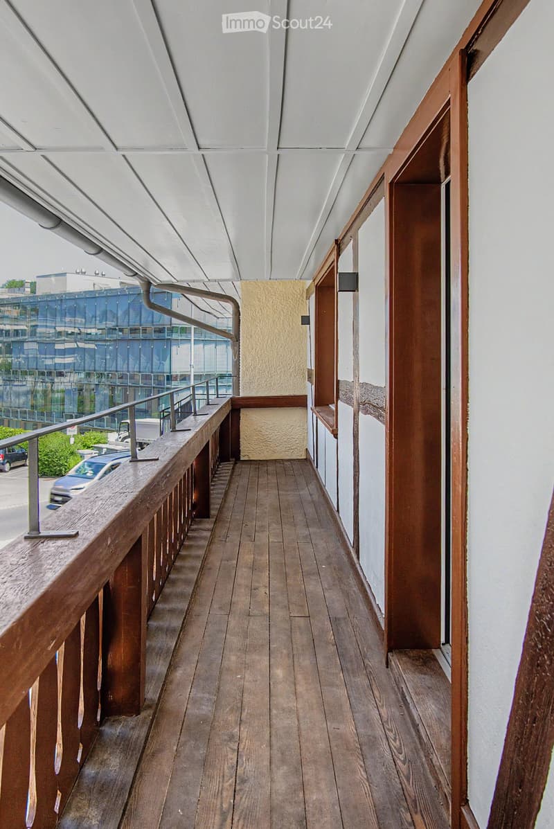 Wooden balcony with metal railing, overlooking a modern building and parking area.