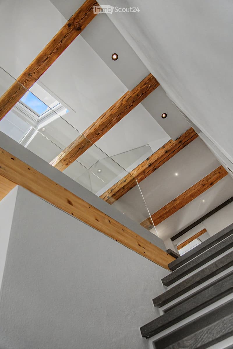 Modern hallway with exposed wooden beams, recessed lighting, and a glass railing leading to an upper level.