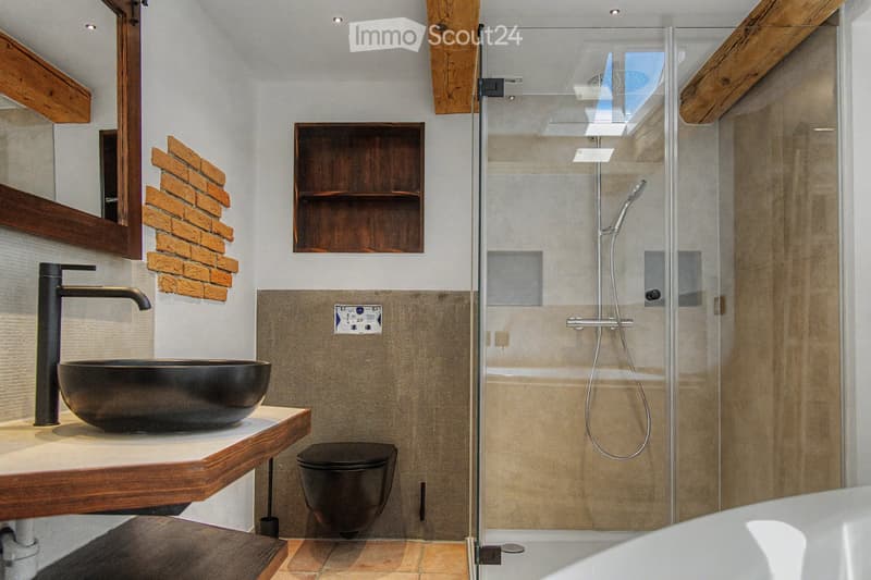 Modern bathroom with a black sink, sleek faucet, wooden shelf, and a glass-enclosed shower area. Features exposed wooden beams and a brick accent wall.