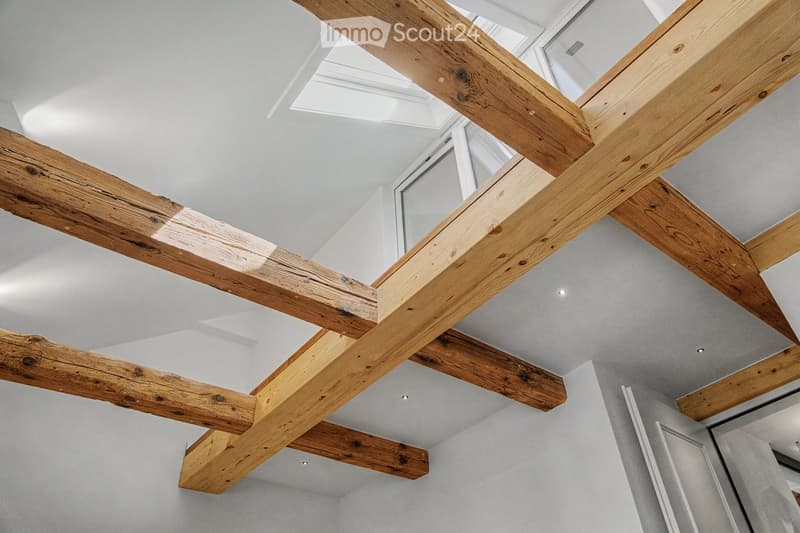 The image shows an interior view of a modern hallway with exposed wooden beams crossing the ceiling. The beams are arranged in a grid pattern, creating a sense of height and openness. The walls are painted a light grey, and the ceiling is white with reces