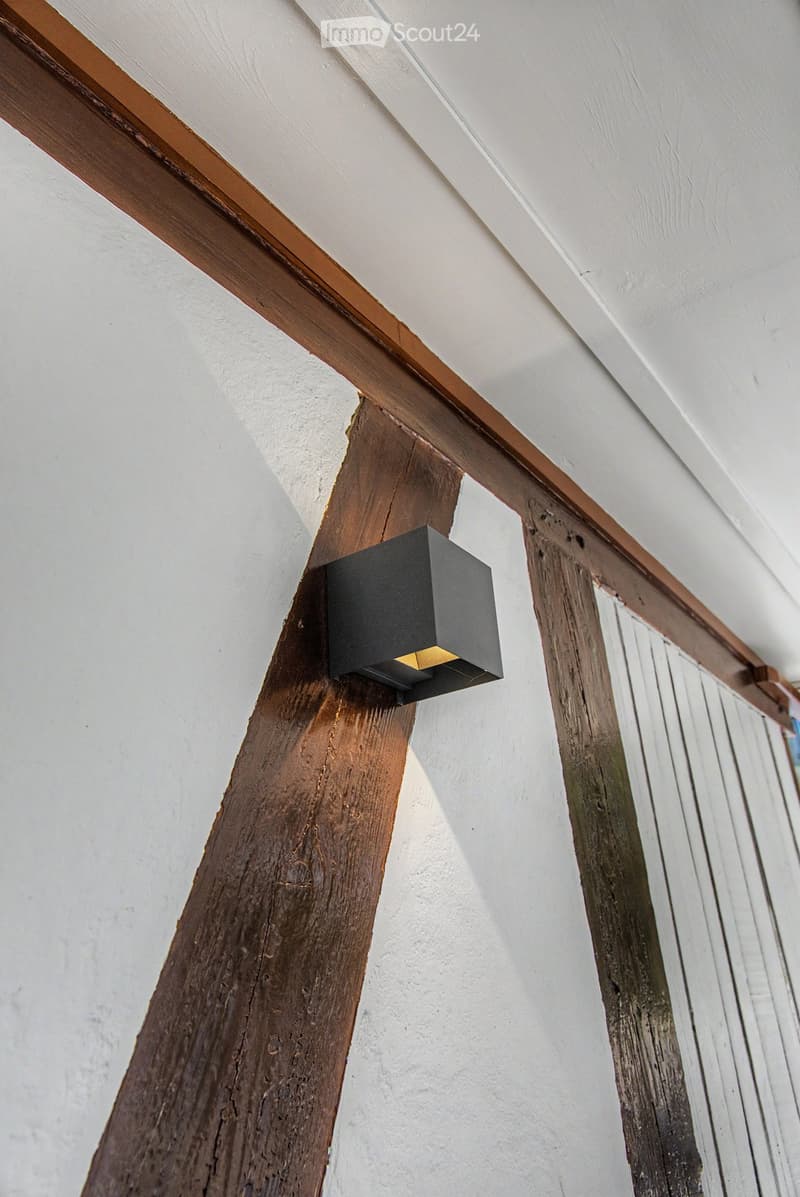 Close-up view of a modern wall-mounted light fixture with a sleek black design, mounted on a wooden beam in a hallway with white walls and exposed wooden beams.
