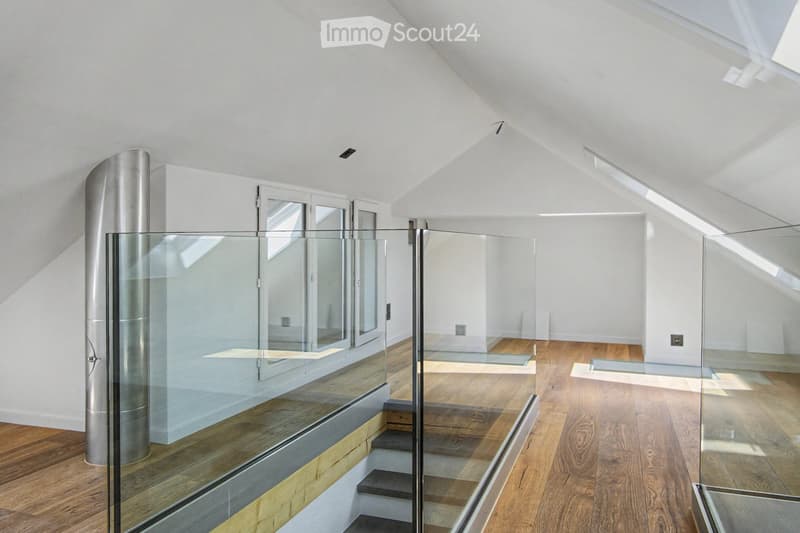 Bright, open loft space with wooden flooring, white walls, and large windows. Features a modern glass staircase with metal railings and a sleek cylindrical column.
