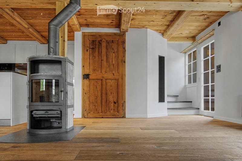 Spacious room with exposed wooden beams, polished wooden floor, large wooden door, and a modern stone fireplace with a chimney pipe.