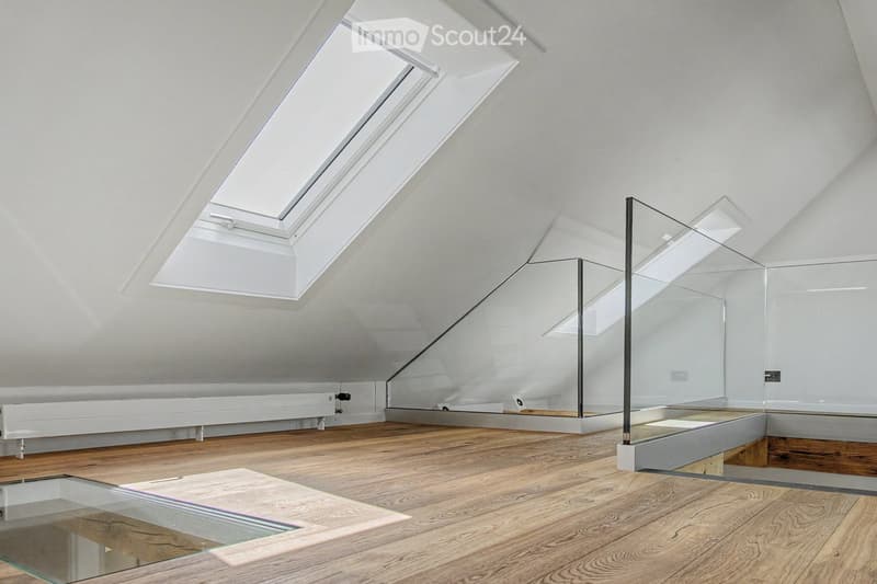 Bright, minimalist attic space with wooden flooring, white walls, and large skylights. Features a glass staircase leading to an upper level.