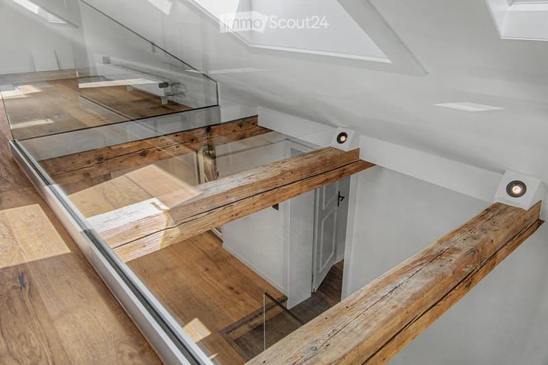 Modern hallway with wooden flooring, glass railing, and exposed wooden beams. The ceiling has skylights, and the walls are white.