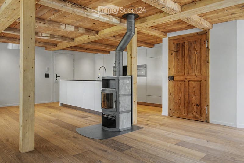 The image shows a modern, open-plan space with a wooden floor and exposed wooden ceiling beams. A wood-burning stove is centrally placed, with a kitchen area visible in the background, featuring white cabinets, a faucet, and an oven. A large wooden door i