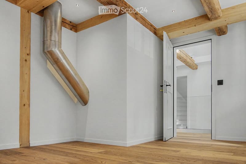Modern hallway with light wooden flooring, white walls, exposed wooden beams, and a stainless steel ducting system. An open door leads to another room with visible wooden flooring and white walls.