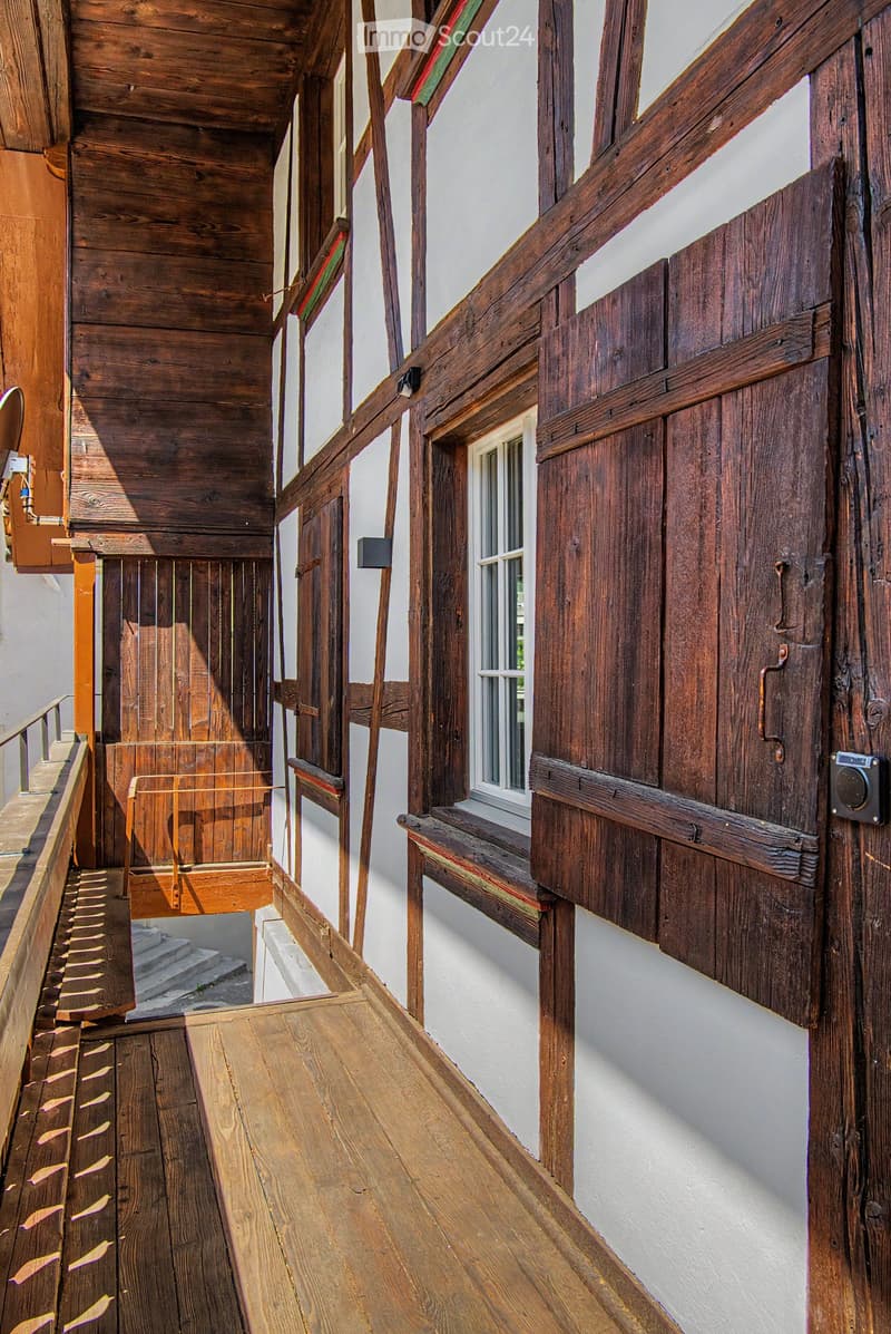 Wooden hallway with exposed beams, white walls, and a small window. Features wooden railings and steps leading to an upper level.